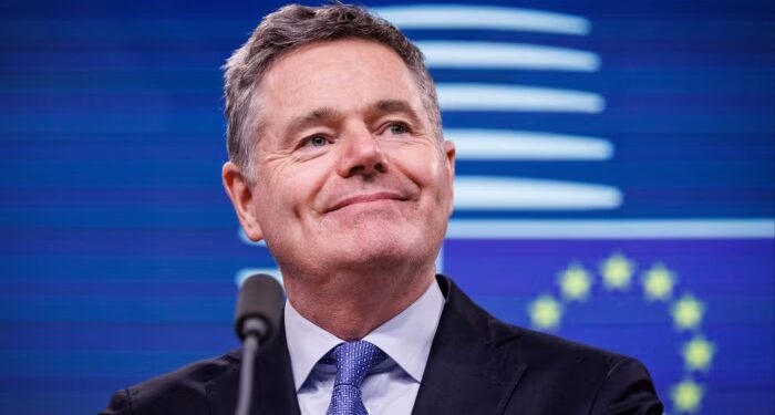 Paschal Donohoe smiles at a podium during a news conference, with the EU flag visible in the background.