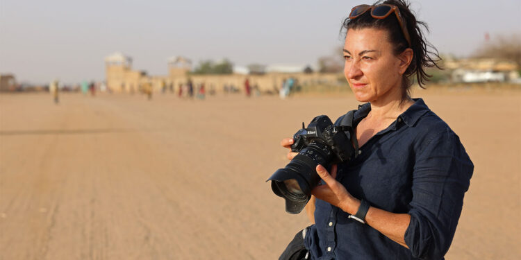 Lynsey Addario: 'There Was Never a World in Which I Would Not Do This Work' A woman wearing a dark shirt and sunglasses on her head stands on a wide, sandy road holding a camera. The background is blurred with people and buildings visible in the distance.