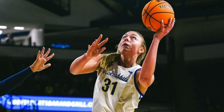 MEYER EARNS PEAK PERFORMER HONOR A women's basketball player, Madison Mathiowetz, is wearing a white uniform and dribbling the basketball. She is guarded by an opposing player wearing a navy blue uniform.