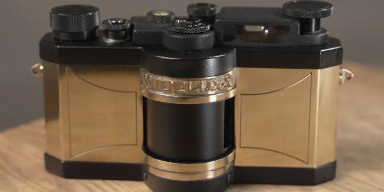The Best Look Yet at Jeff Bridges' WideluxX Camera Revival Project A close-up of a vintage camera with gold and black detailing, labeled "WIDELUX" on a central band, resting on a wooden surface.